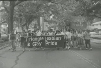 Protestors marching on a street. They hold a banner that says "Triangle Lesbian and Gay Pride."