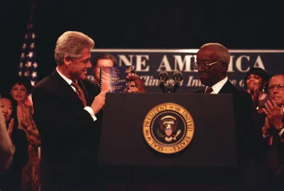 President Clinton stands left holding a book, while John Hope Franklin stands right, holding the same book. They are both wearing suits and are being applauded by onlookers.