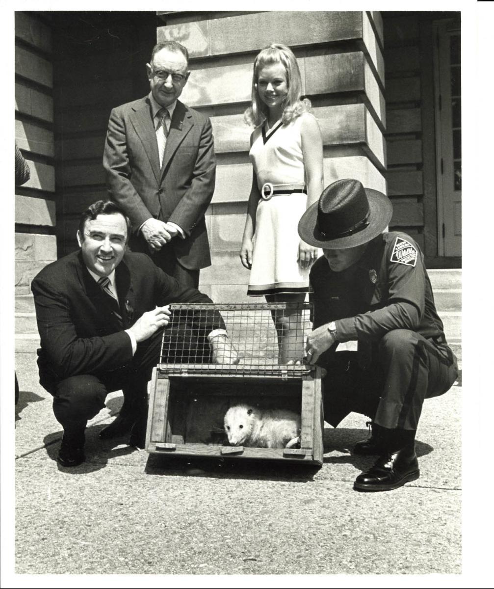A white man in a suit and glasses, a white woman in a dress standing behind two white men, one in a suit the other in a police uniform, kneeling beside a metal cage with Slow Poke the Possum inside. They are in the sunshine outside of the State Capitol building. Black and white photograph.