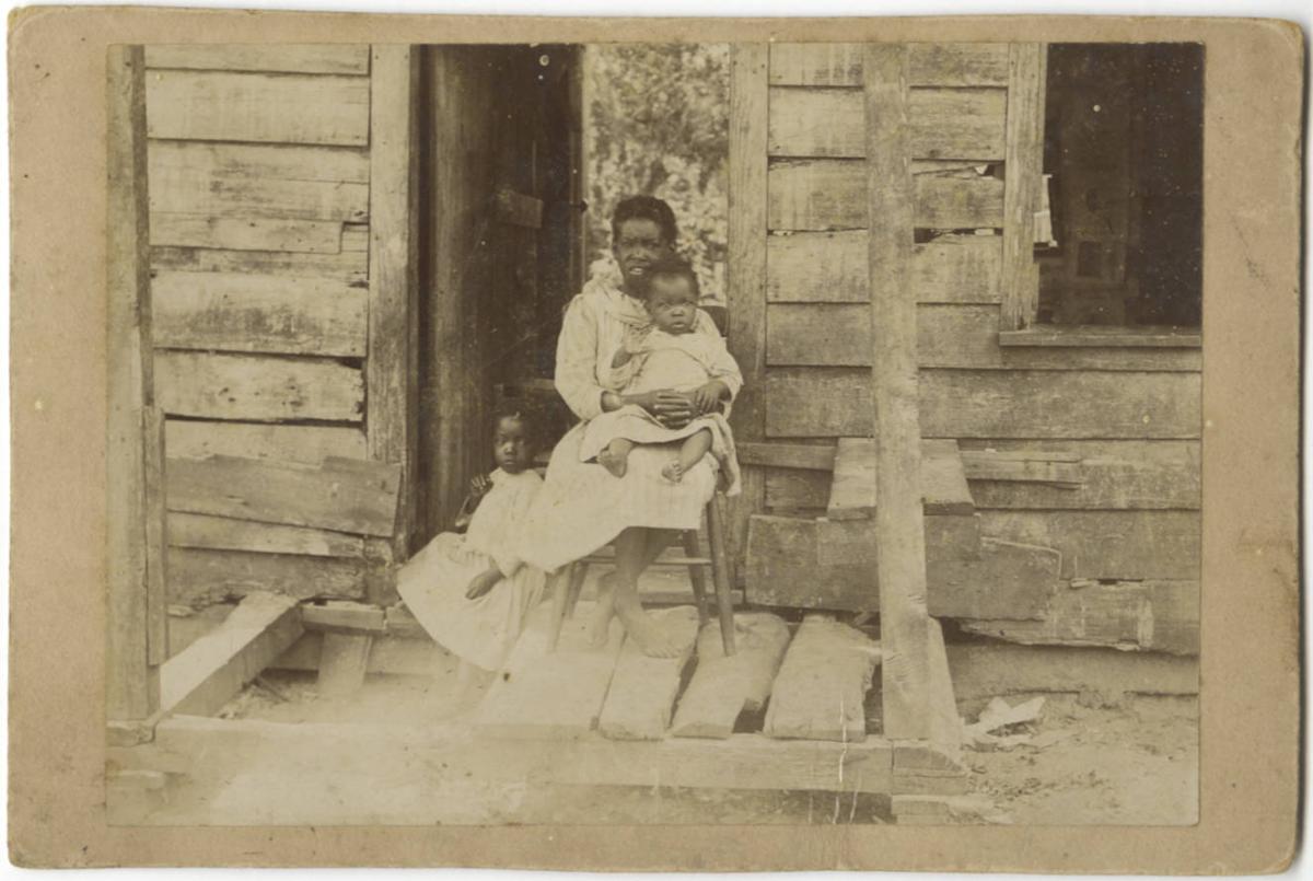 Three children on a porch in front of a wooden-sided home. All three children are black with short hair and white dresses. The oldest child is seated in a chat, holding a toddler in her lap, while another young child sits beside the chair on the porch.