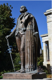 A statue of George Washington. It is bronze and he is depicted wearing a cloak over colonial garb. He has a walking stick in his right hand and a fasces is on his left. Trees, a blue sky, and the Capitol building are in the background.