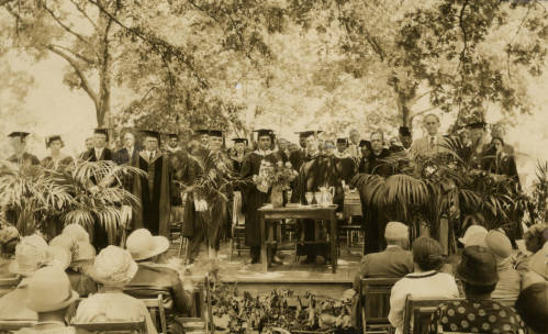 Approximately 30 people wearing caps and gowns on a stage in front of an audience. Dr. David Dallas Jones stands in the middle holding a piece of paper and looking at the camera with a serious look on his face. Black and white photograph.