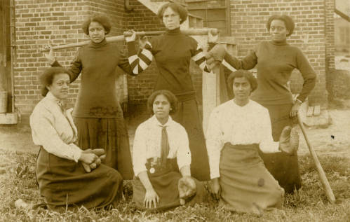 Six young, black women holding baseball equipment. They are all wearing long skirts and long sleeved-shirts. Black and white photograph. 