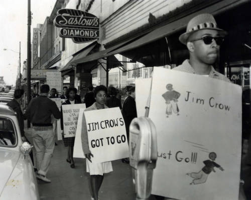 Male and female black protestors wearing signs, walking down a sidewalk under a "Saslow's diamonds" business sign. The protestors signs read: "Jim Crow Must Go!" "Jim Crow's Got to Go!" and "What's right can be done ..." Black and white photograph. 