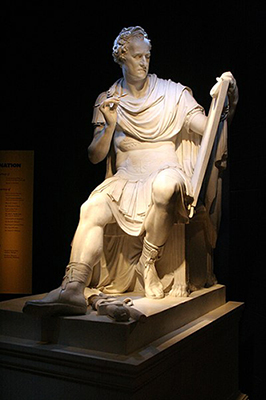 Sculpture of a man dressed as a Roman warroir with a skirt and long cloth around his shoulders. The man is seated and holding up a tablet with his left hand and a pencil like tool in his right hand. 