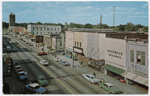 Postcard image of downtown business district on Queen Street in Kinston, N.C. The view is from above the street many cars on the road either driving or parked. There are a few business 
