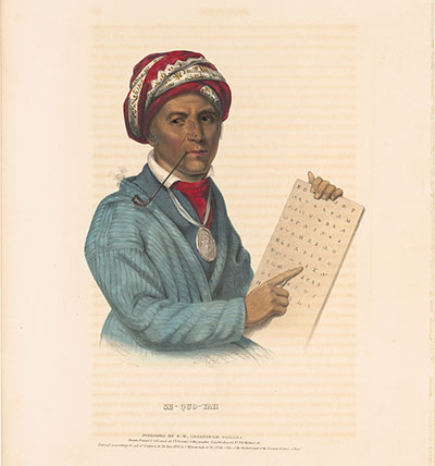 A man with a pipe in his mouth stands holding a tablet with letters he is pointing out. The man is wearing a blue jacket, red head scarf and a medallion around his neck