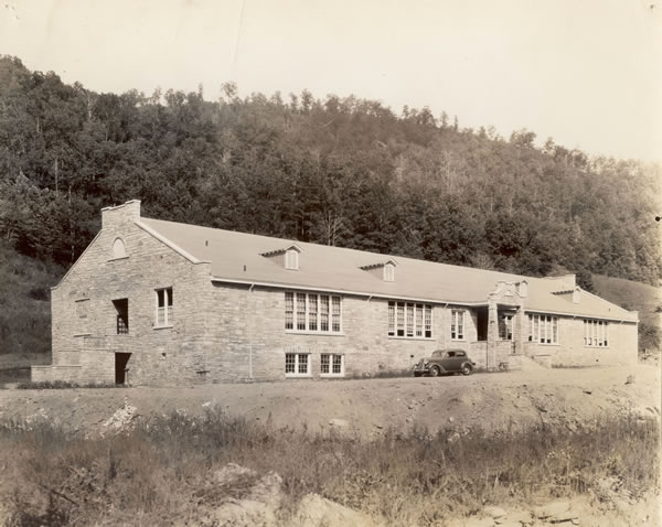Black and white photo of a two story school building. It has several windows. Behind the building are large trees and in front there is an old automobile 
