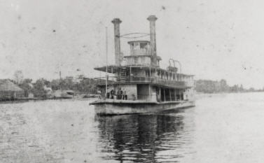 A boat on water close to the shore. The boat has two large chimney-like towers and three levels. The photo is black and white.
