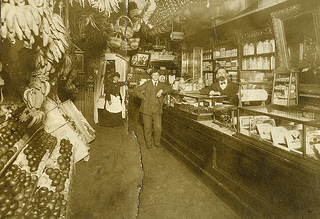 Two men in suits stand at a counter. A woman in a long black dress and white apron stands behind them. Produce are on shelves on the left of the picture.
