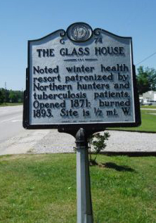 Large metallic marker that reads: "THE GLASS HOUSE, Noted winter health resort patronized by Northern hunters and tuberculosis patients. Opened 1871; burned 1893. Site is 1/2 mi. W."