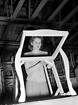A person stands assembling a wooden chair in a building. 