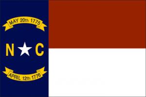 The flag of North Carolina. On the right side is a large horizontal red panel above a horizontal white panel. On the left side is a long, narrow blue panel. Text in the blue panel reads May 20, 1775, NC, April 12, 1776. A white star appears between the letters N-C.