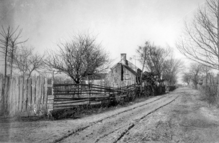 A dirt path with tracks rutted wheel tracks down it.  The path goes along a wooden fence. Behind the fence is a run-down wooden building with a chimney.