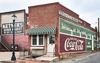 A brick building. A striped awning is over the door and windows into the building. "Ketner's Super Market" and a Coca Cola ad are painted on the building walls.