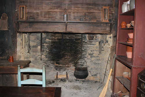 A colonel kitchen with a caldron near a log of firewood. There are two tables and a large shelf with bowls, cups and plates.  Kitchen at Allen House, Alamance County, N.C., where John and Rachel Allen lived with their family in the late 1700s.