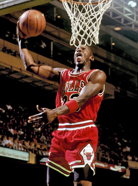 Michael Jordan dunking a basketball. He wearing his red Chicago Bulls jersey and shorts. He is mid-air and there are many people in the stadium.