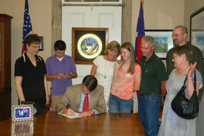 A group of people standing behind a man signing a document. While in a government building with the state seal, and both American and state flag  in the background.