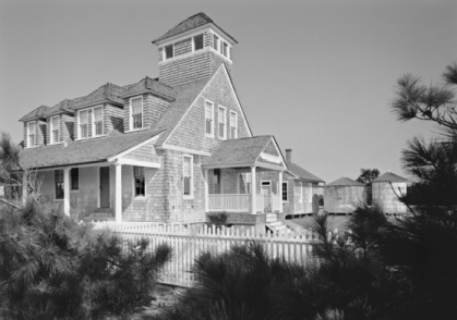 A large wooden building. There are bushes and trees in the foreground. Black and white photo