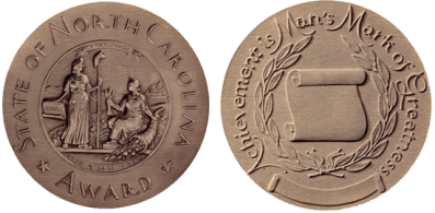 Image of the front and back of the North Carolina Award medal. The medal is gold in color and composition and has raised images and text. The front reads "State of North Carolina Award" with the state seal, an image of two women in robes representing Liberty and Plenty, one holding a capped pole and one holding heads of grain with a cornucopia. The back of the medal says "Achievement is Man's Mark of Greatness" and has an unfurling scroll surrounded by leafy branches.