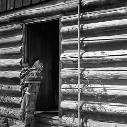 A woman with child wrapped on her back stands in front of a log building.