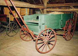 A green wooden wagon with red wooden wheels. There is what appears to be a shelf that covers a compartment in the bottom of the wagon. There is a pile of hay in the wagon, as well. 