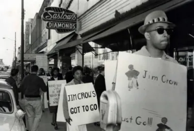 Male and female black protestors wearing signs, walking down a sidewalk under a "Saslow's diamonds" business sign. The protestors signs read: "Jim Crow Must Go!" "Jim Crow's Got to Go!" and "What's right can be done ..." Black and white photograph. 