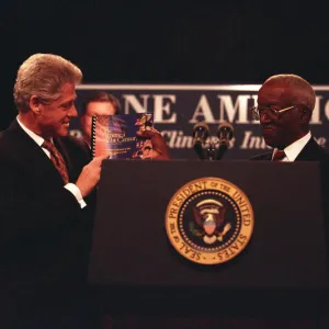 President Clinton stands left holding a book, while John Hope Franklin stands right, holding the same book. They are both wearing suits and are being applauded by onlookers.