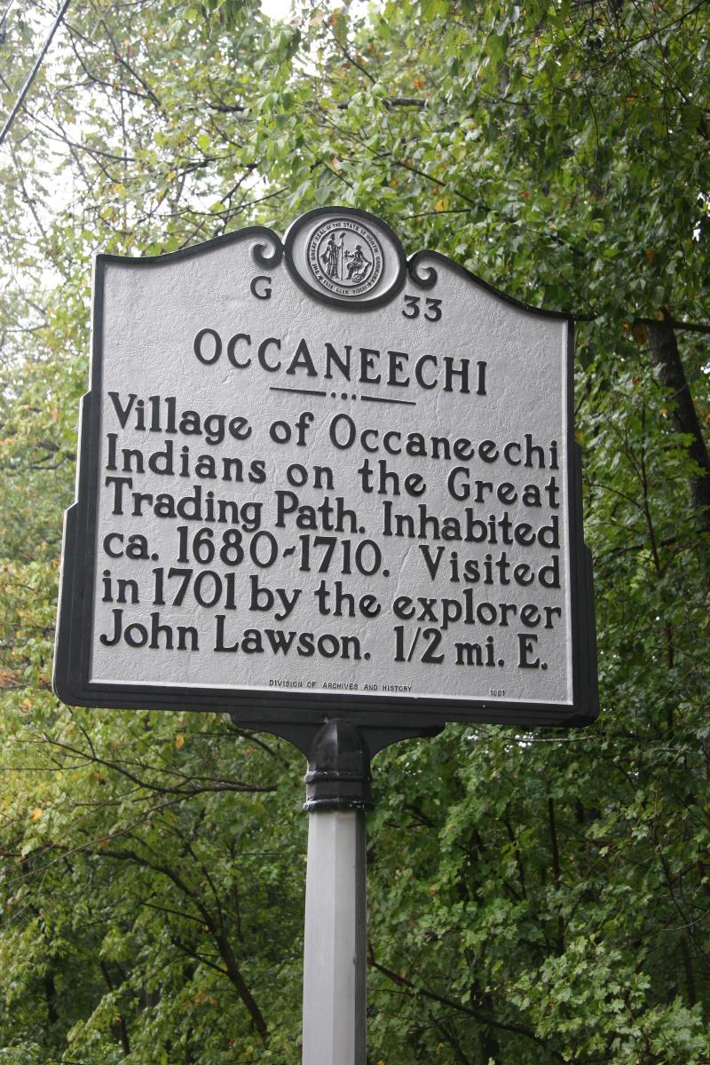 Large metallic marker that reads: "OCCANEECHI, Village of Occaneechi Indians on the Great Trading Path. Inhabited ca. 1680-1710. Visited in 1701 by the explorer John Lawson. 1/2 mi. E."
