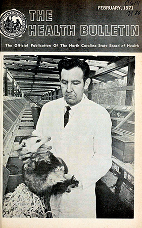 The cover of The Health Bulletin, February 1971. Dr. Jurgelsky removes an opossum from a nest box in the building developed at the National Institute of Environmental Health Sciences, Research Triangle Park,  to house the opossum breeding colony. Individual cages and nest boxes are seen to either side. In order to avoid a painful, and often severe bite on the hand, the animals must be restrained by quickly and firmly grasping the nape of the neck.