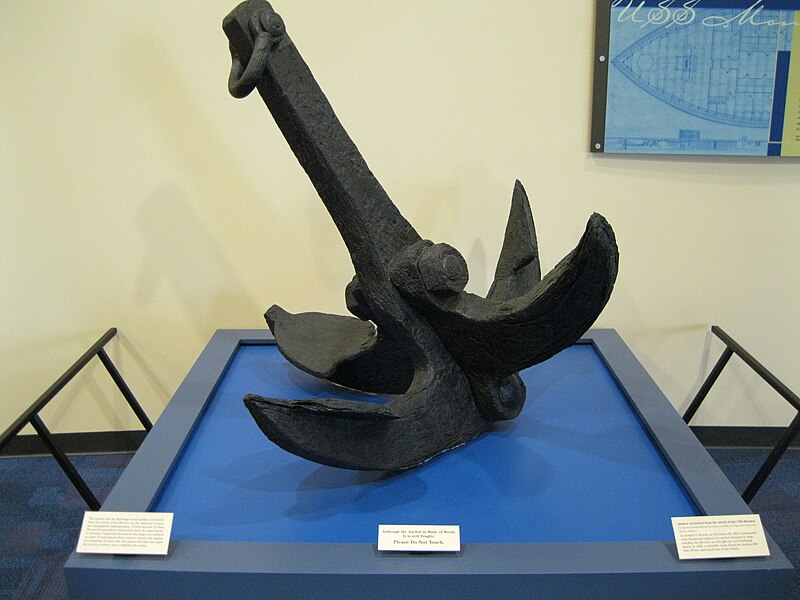 A large, metal anchor sits on a display table. It has four large hooks that stick out from each side. 