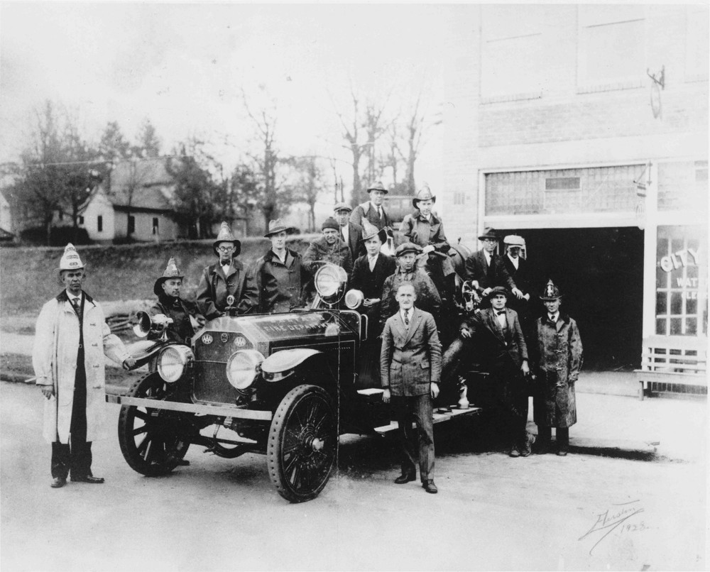 Black and white photograph of firemen in and around a firetruck in front of a firehouse, circa 1913-1930
