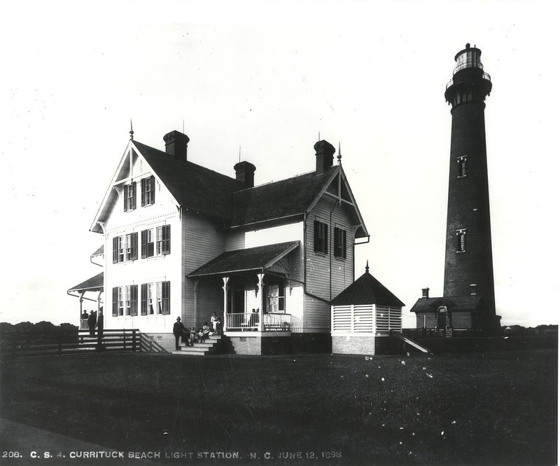 A lightkeeper's house sits beside the brick Currituck lighthouse. There are people on the porch of the house. Black and white photo.