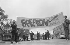 Black and white photograph of people walking down the street, as if in protest. Two men wearing suits are at the front carrying a large banner that reads "FREEDOM FOR ALL PEOPLE!"