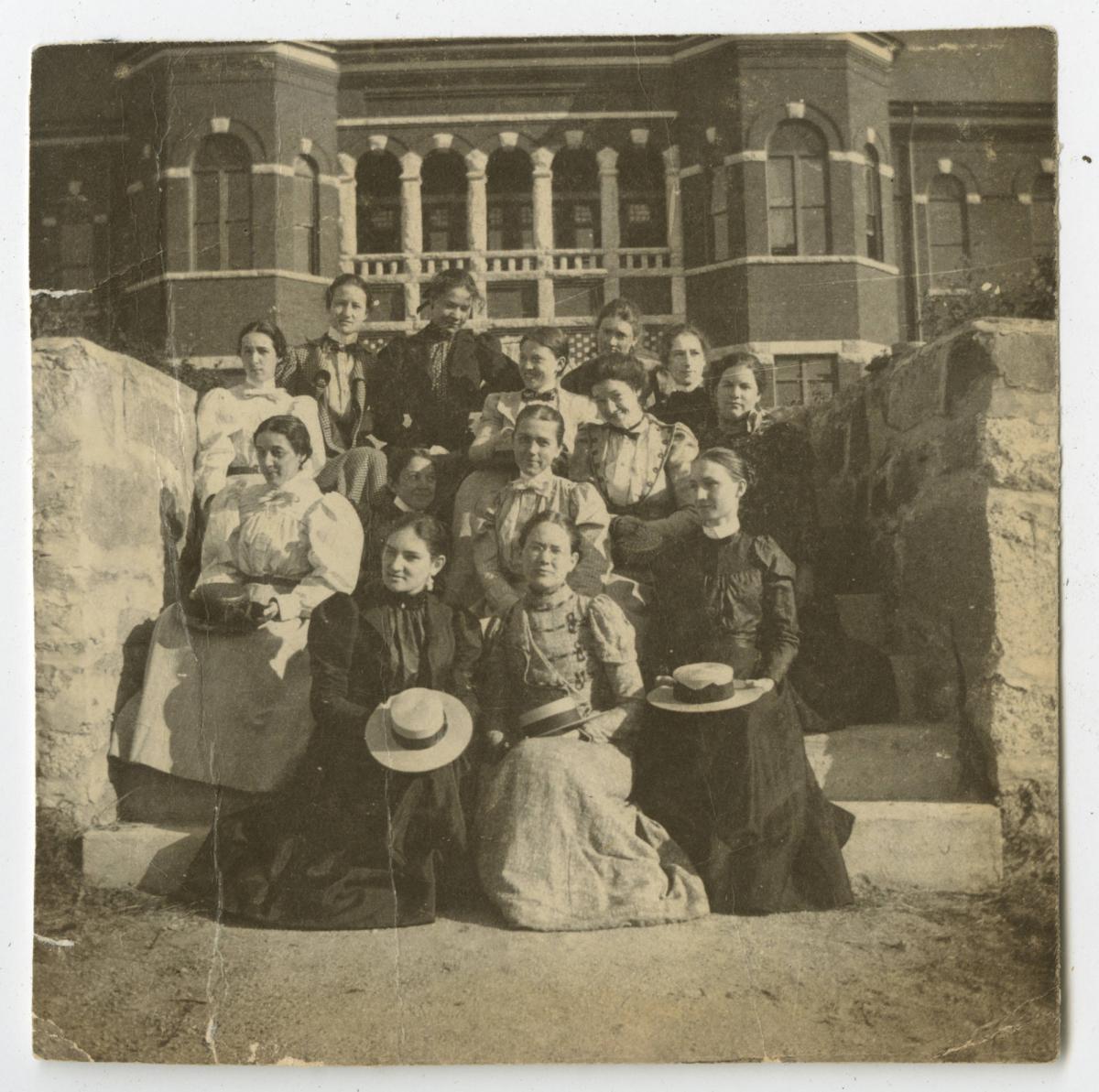 Thirteen students sit on the steps of a campus building for a group photo.
