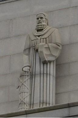 A statue on the ledge of a building exterior. It depicts a robed figure with a styled beard. He is holding his left hand to his chest and has his right arm resting on a fasces. 