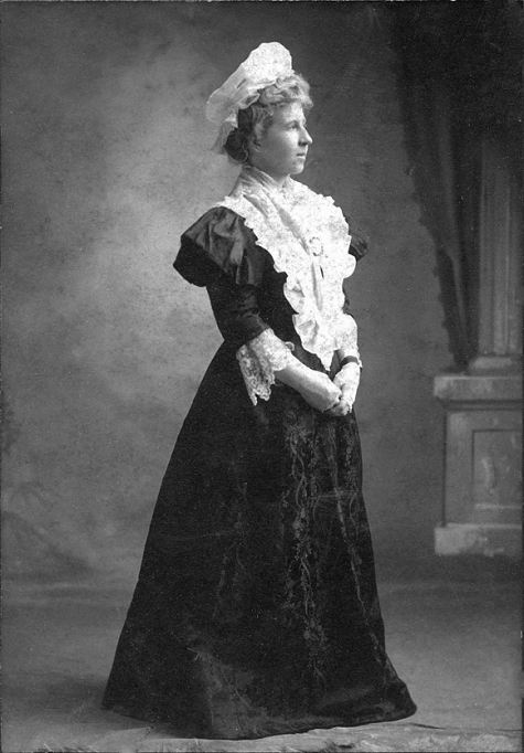 Black and white photo of woman wearing long black dress, white lace collar, and white hat.  
