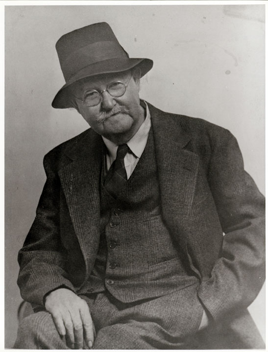 Black and white photo of a white man with round metal glasses and a hat sitting for a portrait. He is wearing a 3-piece-suit and a tie. One hand is in his pocket and he has a slight grin. 