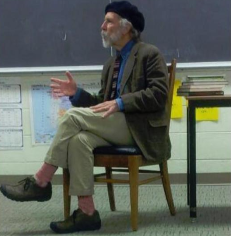 Bathanti speaking in a classroom. He is wearing a beret, a tweed jacket, and khakis. He is older with a white beard. 