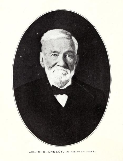 A black and white photograph of Creecy at age 94. He is older. He has short, white, styled hair and a beard. He is wearing a suit.