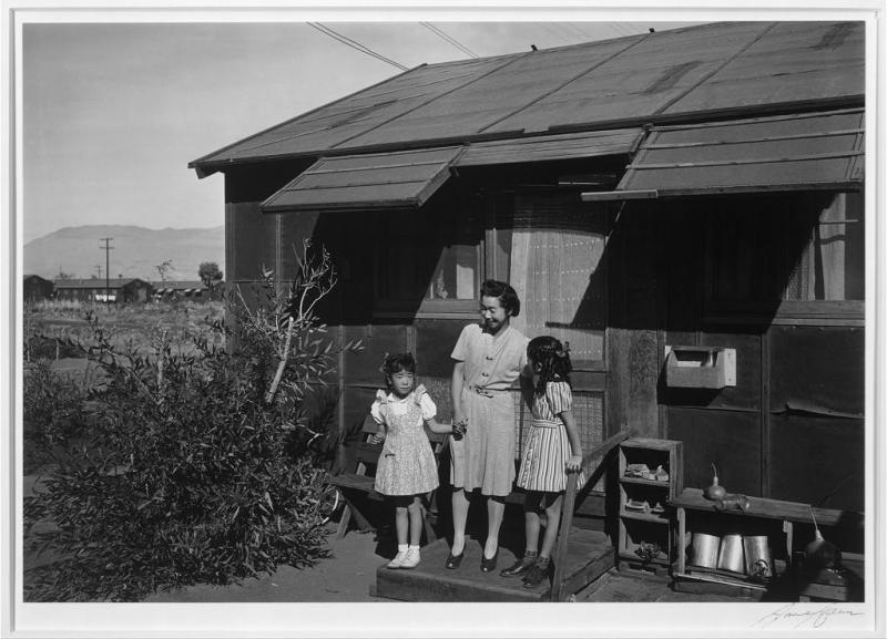 Mrs. Naguchi and two children, Manzanar Relocation Center An Asian woman stands in front of her relocation house with her two daughters.