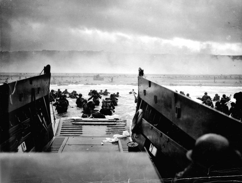 A landing boat shores on a beach. Soldiers wade through water in military equipment. 