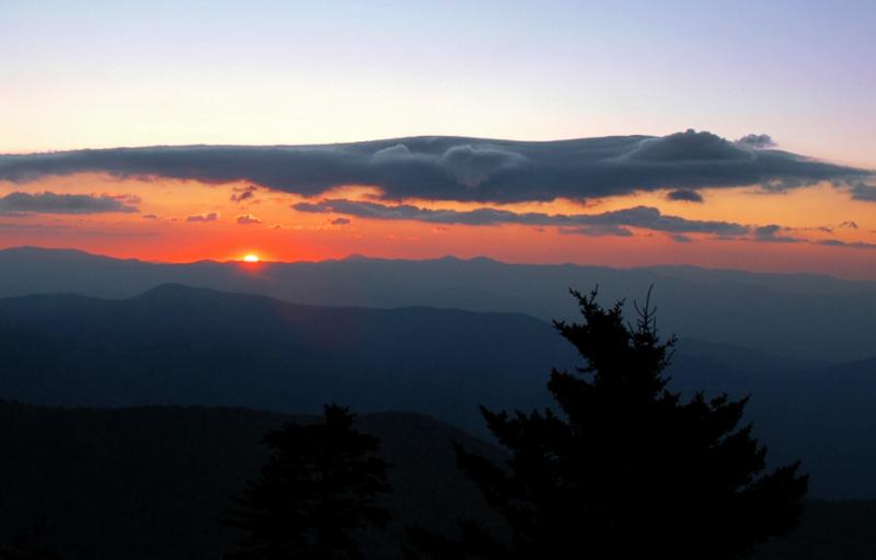 A sunrise coming over a mountain top. Orange skies and blue clouds depicted.