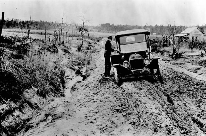 A vintage automobile stuck in mud on a road. The road is cut through a hill, with banks on each side. A man is trying to get the car out of the mud. Their is a cabin in the background. Black and white. 