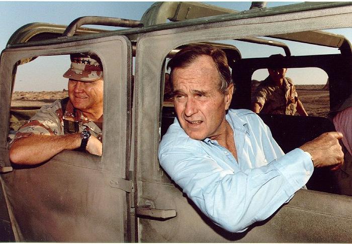 George HW Bush and Norman Schwarzkopf in a jeep in the desert. Both have concerned expressions and are squinting from the sun. 