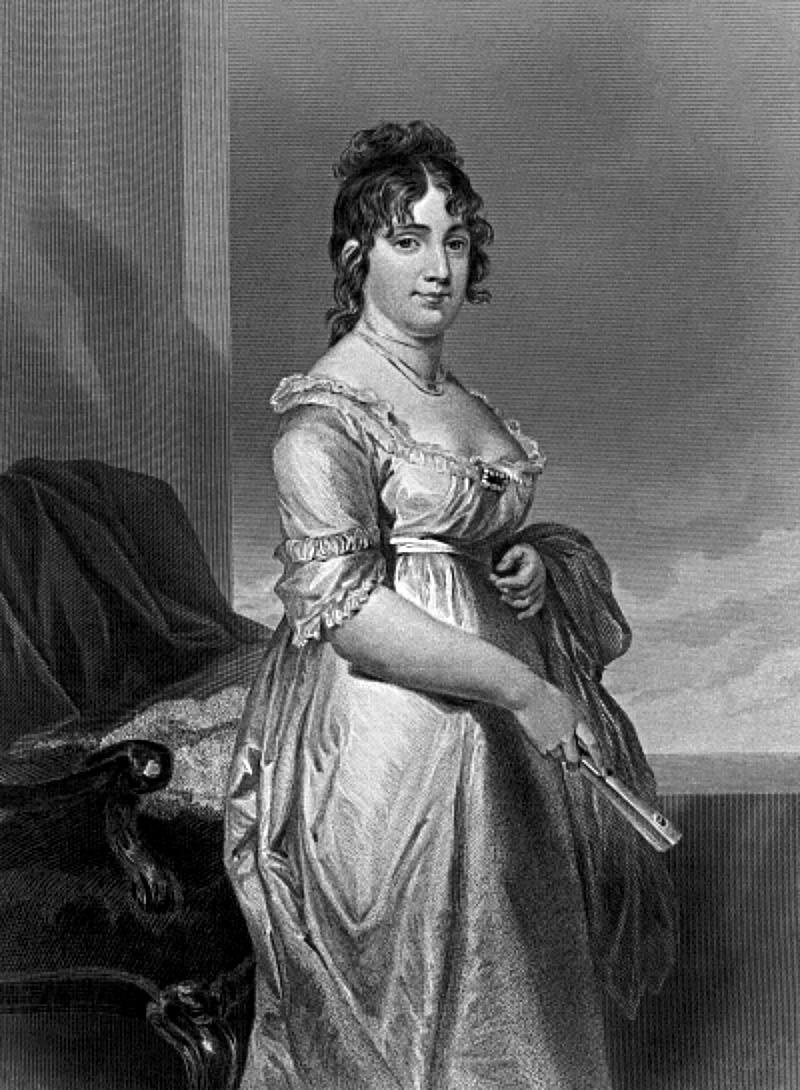 Dolley Madison. Black and white. She is standing beside an armchair wearing a light dress with her hair up. She is holding a hand fan. 
