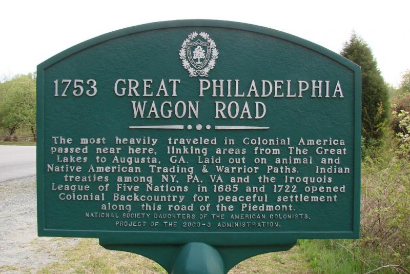 Great Wagon Road marker. It is green and surrounded by brush and there is a road in the background. 