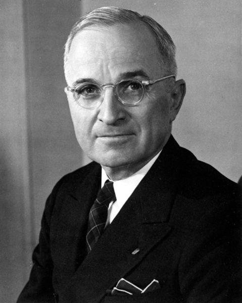 Photo of President Truman. He has glasses and short hair. He is smiling while wearing a suit.