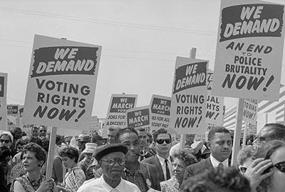 This photograph features black and white people holding signs at the March on Washington, 1963. One sign reads: We demand Voting Rights and is held by a white woman. Another sign reads: We demand an end to police brutality now and is held by a black man.
