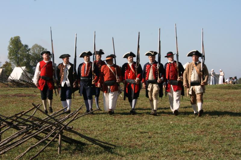 Nine reenactor soldiers in a formation. They are wearing apparel of the colonial military, with tricorn hats and buttoned coats. It is sunny.